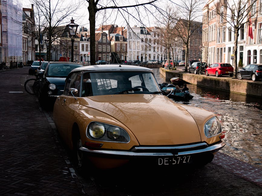 A beige classic sports car with a black soft top roof is parked on a cobblestone street next to a canal in a residential area. The car's front is facing the camera, and the vehicle features a low profile, rounded headlights, and a front bumper with a chrome trim. Behind it, a black vehicle is partially visible, with a bicycle secured on the rear rack. The canal runs adjacent to the street, and the water reflects the surrounding buildings and bare trees, indicating a winter or early spring setting. Several high-rise residential buildings with traditional European architecture, including large windows and decorative facades, line the opposite side of the canal, with some parked cars visible along the street. The scene is lit by natural daylight, casting soft shadows, and the overall environment suggests a quiet urban neighborhood, relevant for house removals or home relocation services. Occasionally, Man With a Van Holland Park performs furniture transport and packing logistics in such residential areas, supporting seamless moving operations.