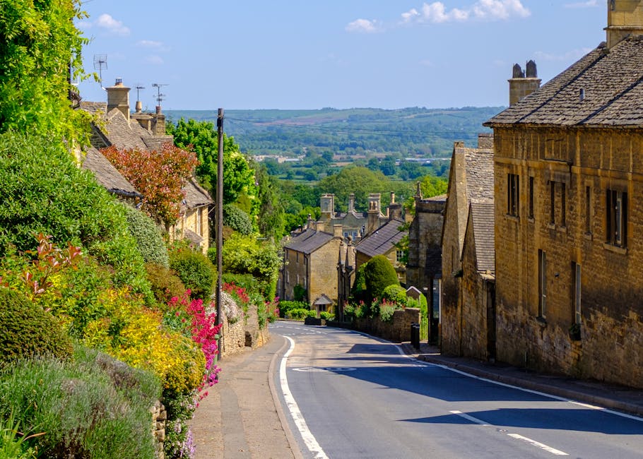 A narrow residential street in a historic village, showing stone-built houses with slate roofs on either side, surrounded by lush green trees and flowering shrubs. The road gently slopes downward, wit