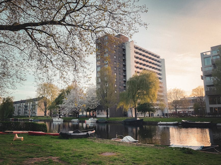 A view of a residential area near Holland Park with a canal in the foreground, showing several small boats moored along the grassy bank. Large trees with budding leaves and blossoms surround the canal, their branches extending overhead. In the background, there is a tall, modern high-rise apartment building made of concrete and glass, alongside other mid-rise buildings with contemporary designs. The scene is captured during daylight with soft, natural lighting indicating early morning or late afternoon. In the vicinity, there is no visible activity related to house removals or moving processes; the focus is on the urban landscape and natural surroundings, which provide context for the locality served by Man With a Van Holland Park's removals services at [PAGE_TITLE].