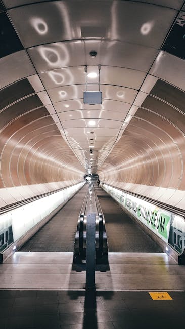 A long, enclosed underground corridor at Holland Park station featuring curved, metallic walls and a ceiling with circular lighting fixtures. An ascending escalator is centered in the image, with a handrail on each side, leading towards the surface exit. The floor is made of tiles, and an illuminated green and white sign along the right wall displays station information. The environment appears clean and modern, with the lighting creating a bright, welcoming atmosphere suitable for passengers during home relocation or moving logistics planning, as managed by Man With a Van Holland Park. This scene relates to efficient transportation and the final stages of furniture transport or packing and moving preparations.