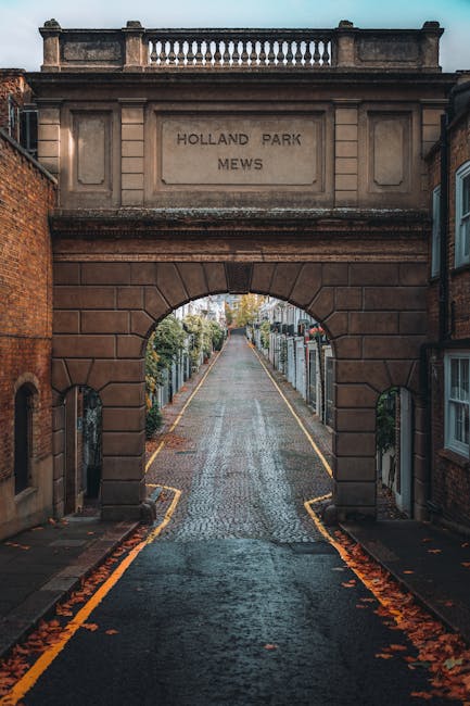 A narrow, cobbled street viewed through an arched stone gateway inscribed with 'HOLLAND PARK MEWS', with residential buildings on either side. Inside the gateway, several cardboard boxes and wrapped furniture pieces are visible on the pavement, indicating a house removal or moving process. Visible in the scene is a hand truck or trolley positioned near the boxes, and a person’s hand or arm may be assisting with the loading or unloading activities. The street is lined with fences and greenery, with soft natural lighting suggesting early morning or late afternoon. The environment reflects a quiet residential area suitable for professional moving services by Man With a Van Holland Park, with materials prepared for home relocation or furniture transport within the neighbourhood.