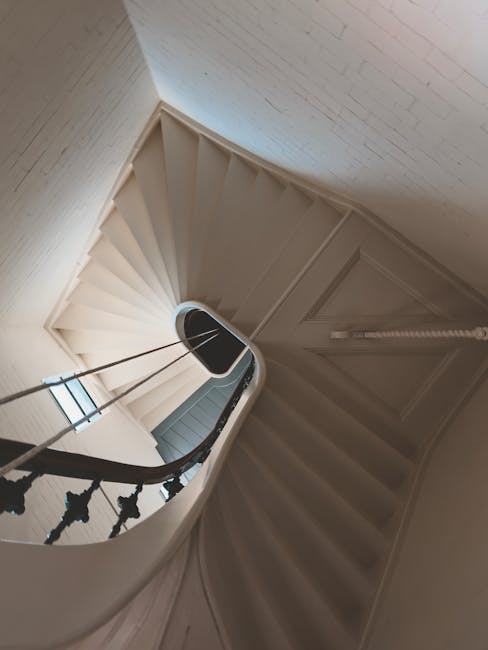 A narrow, spiral staircase inside a residential property, with white-painted wooden banisters and black decorative spindles. The stairs are made of light-colored wood or laminate and are closely spaced due to the staircase's tight turn. Natural light enters through a window at the top of the staircase, illuminating the area. The staircase appears to be used during a house move or furniture transport process, with the setting indicating a home relocation environment. The image is taken from a top-down perspective, capturing the curved steps descending towards an enclosed lower level. The surrounding walls are painted light, and the overall environment is clean and tidy, supporting professional removal activities by a company such as Man With a Van Holland Park.