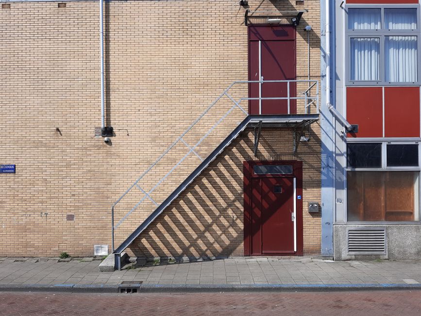 A metal fire escape staircase with a shadow cast on a beige brick exterior wall of a residential building. The staircase has a railing on both sides and leads up to a red door on the first level. Below the staircase, there is a small passageway with a red door that appears to serve as an entrance or access point to the building. Adjacent to the staircase on the right, there is a window with a red and gray panelled exterior, as well as a vent and a mailbox mounted on the wall. The scene is illuminated by daylight, with a sidewalk and a drainage grate visible at street level, indicating urban surroundings consistent with house removal or relocation logistics. This setting exemplifies common challenges faced during home relocations involving narrow staircases and access constraints, which Man With a Van Holland Park manages as part of their removals services.